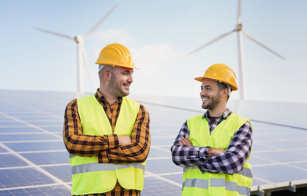 Worker men at solar power station - Solar panels with wind turbines in background